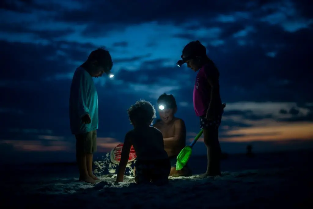 Four kids in the sand on the seaside with batterie lamps on their foreheads, in the dark, looking at the sand, two of them are sitting and the other two are standing, all engaged play of digging the sand and looking for something