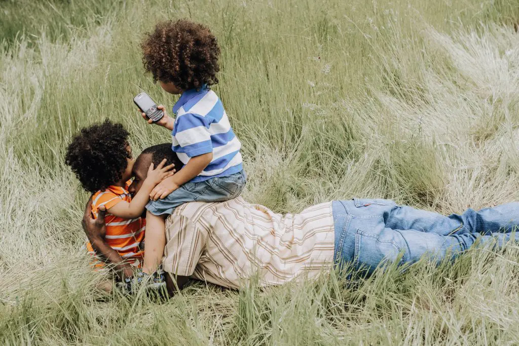 a family of three in the grass, a father laying on the stomach and one child siting on his shoulders and tapping on the mobile, while the other one is in his arms and they're hugging. Both kids have frizzy African-American hair