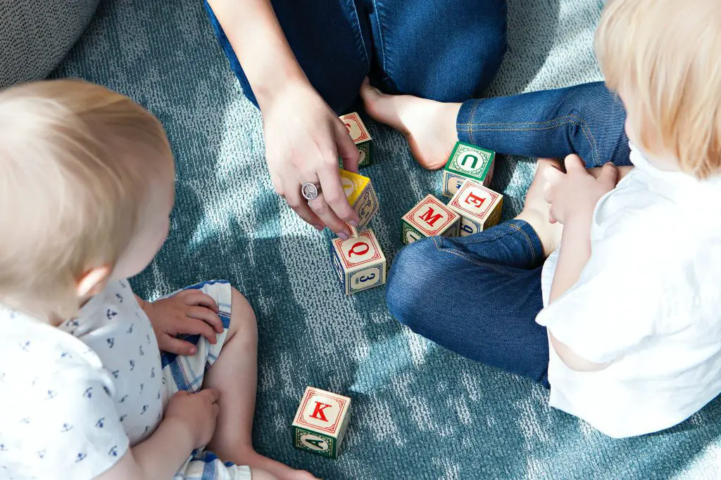 Two toddlers sitting on the flor while a woman is teaching them letters by rolling the dice with letters on them
