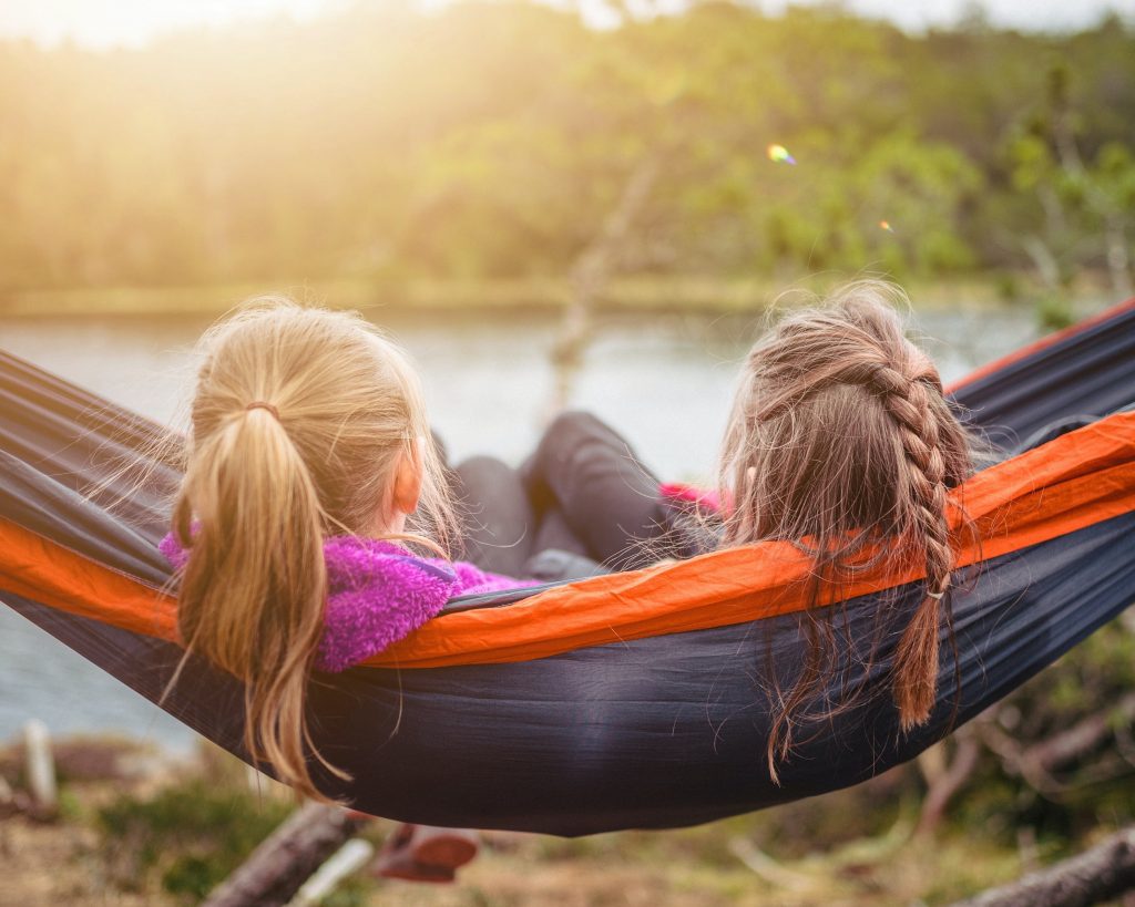 a landscape of two little girls with pony tail and braids are peacefully sitting in a swing in nature and looking at the water