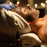 Close-up of peaceful and calm young woman getting skin treatment with clay mask. Female covered with white towel. Joy and relax at cosmetologist. Beauty and spa day concept