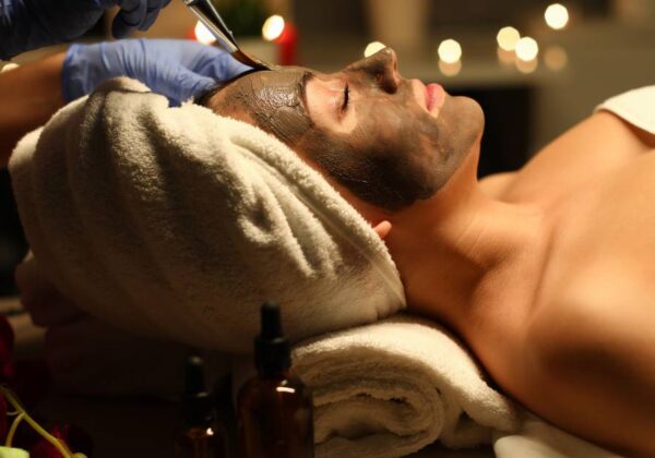 Close-up of peaceful and calm young woman getting skin treatment with clay mask. Female covered with white towel. Joy and relax at cosmetologist. Beauty and spa day concept