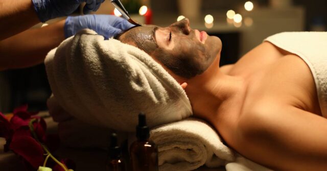 Close-up of peaceful and calm young woman getting skin treatment with clay mask. Female covered with white towel. Joy and relax at cosmetologist. Beauty and spa day concept